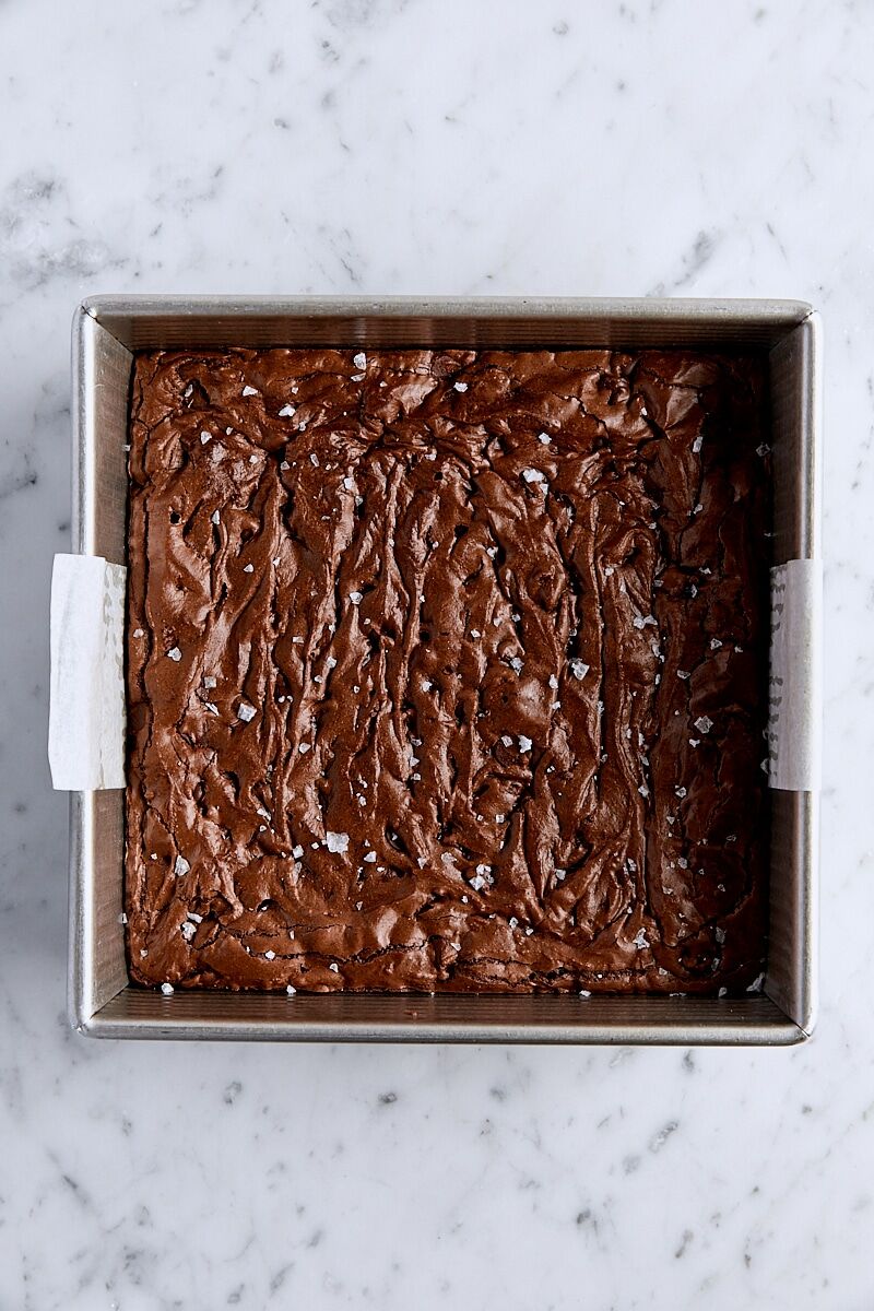 Baked brownies topped with flaky salt in parchment-lined square pan
