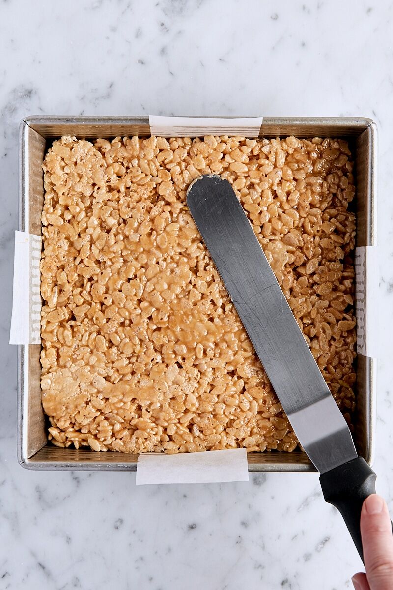 Rice krispie mixture pressed into parchment-lined pan with metal spatula