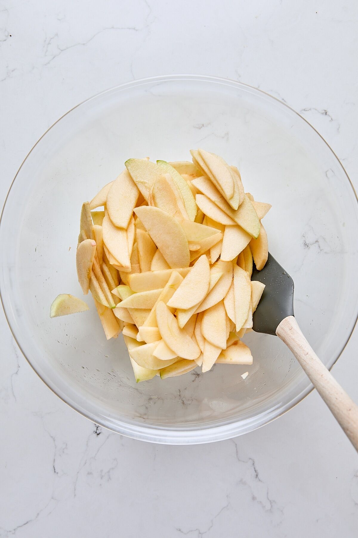 Sliced apples tossed with lemon juice and vanilla in glass bowl with spatula